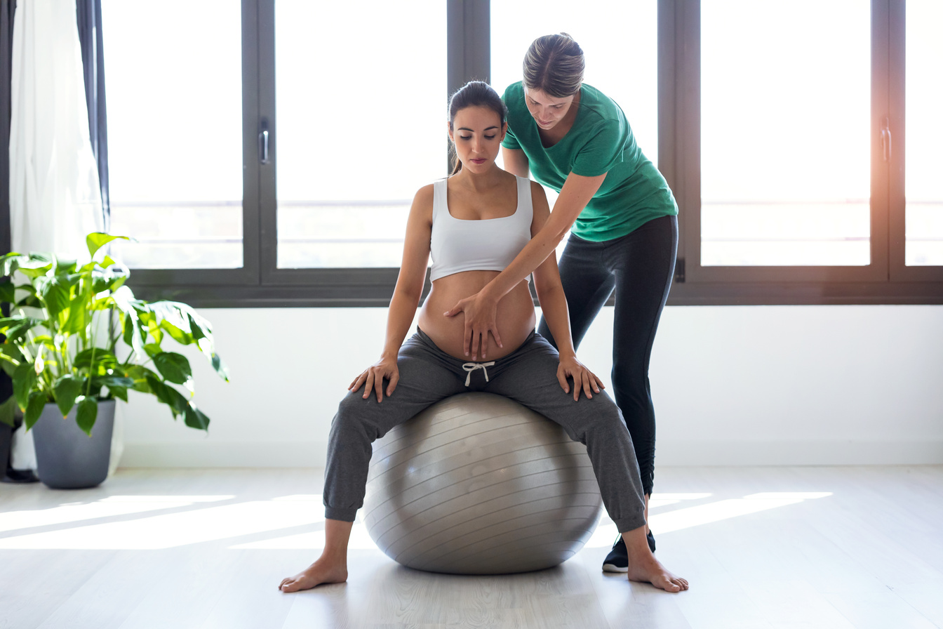 Physiotherapist helping to beautiful pregnant woman for doing pilates exercises with ball preparing for childbirth.