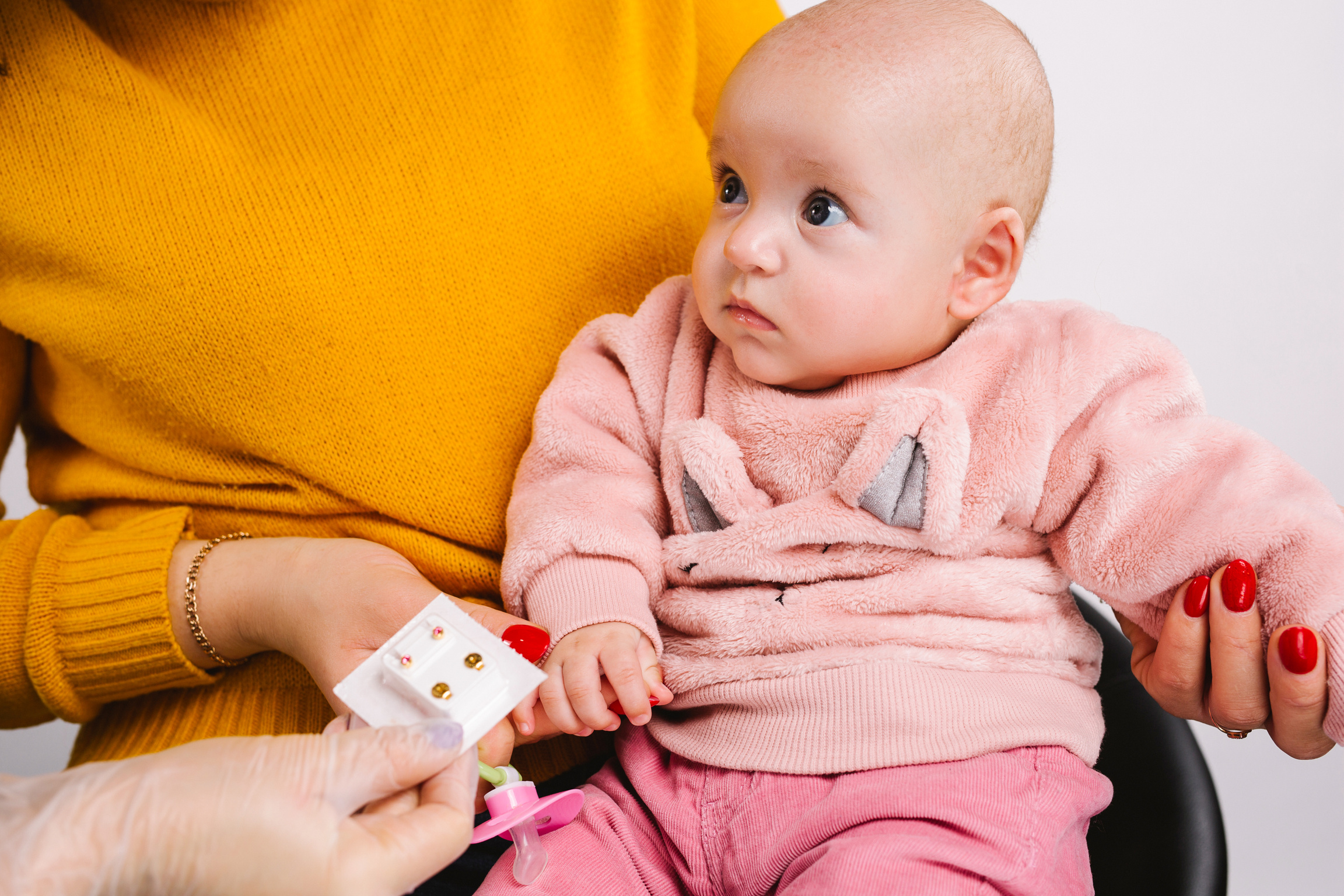 Cropped photo of a cute baby in mummy arms choosing golden small medical earrings for ear piercing. White background.