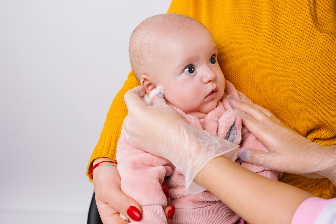 Enlarged photo of a little cute baby girl whose doctor's hands rub her ear in preparation for the ear piercing, beauty procedure.